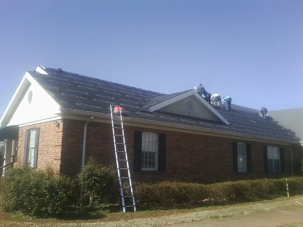 Crew tearing shingles off a brick ranch roof with a ladder set — Emergency Roof Repair work by our crew in Eure