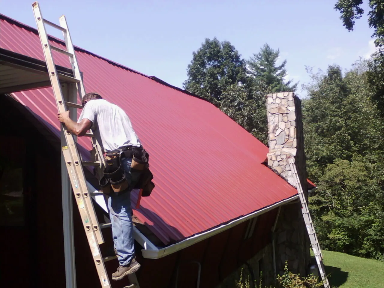 Roofer on a ladder beside a red metal roof and stone chimney — Soffit & Fascia Repair by Roofing Brothers in Eure